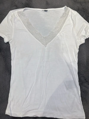 Vit t-shirt med spetsdetalj - Vit t-shirt med djup v-ringning och spets längs urringningen. Kortärmad och i mjukt bomullsmaterial. Perfekt för en enkel och stilren look.