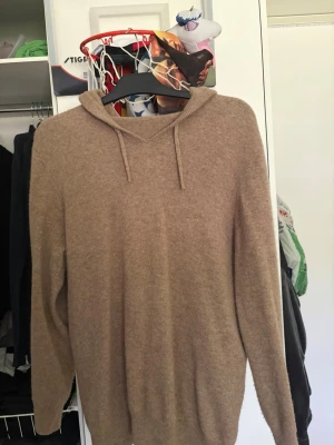 Beige stickad hoodie med luva - Mysig beige stickad hoodie med luva och dragsko. Tröjan har långa ärmar och ribbade muddar vid ärmslut och nederkant. Perfekt för dig som gillar en avslappnad och stilren look. Materialet känns mjukt och fluffigt, troligen ull- eller kashmirblandning.