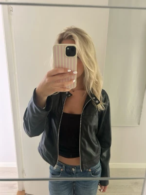 Svart skinnjacka med dragkedja - Snygg svart skinnjacka med silverfärgad dragkedja framtill och detaljer vid kragen. Jackan har en rak passform och långa ärmar, perfekt för en cool och stilren look.