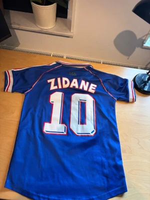 Frankrike Zidane tröja VM 98 Adidas - Blå fotbollströja från Adidas med Zidane och nummer 10 på ryggen. Officiell Frankrike VM 98-design med vita och röda detaljer, broderad tupp och FIFA World Cup-märke på bröstet. Klassisk krage och ränder på ärmarna. Perfekt för fotbollsfans!