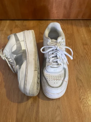Vita Nike Air Force 1 sneakers - Klassiska vita Nike Air Force 1 sneakers med lågt skaft och perforerade detaljer på tån. Skorna har vit sula och snörning samt den ikoniska swoosh-loggan på sidan. Tillverkade i läder och textil för en clean och tidlös look.