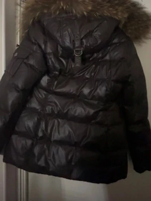 Svart dunjacka med pälshuva Hollies - Snygg svart dunjacka från Hollies med stor huva kantad av fuskpäls. Jackan har quiltad design, dragkedja och dekorativt mönstrat innerfoder. Perfekt för kalla vinterdagar och riktigt varm tack vare dunfyllningen.