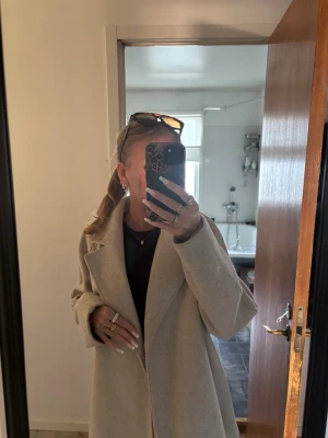 Beige lång kappa med bred krage - Snygg beige kappa med rak passform och bred krage. Kappan har en stilren design utan synliga knappar och är perfekt för dig som gillar en clean och trendig look. Materialet ser ut att vara ullblandning och kappan är helfodrad för extra komfort. Har tappat bort snöret till kappan men den ser fortfarande lika bra ut.