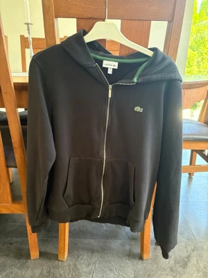 Svart zip-hoodie från Lacoste - Säljer en svart hoodie från Lacoste med dragkedja framtill och klassisk krokodil-logga på bröstet. Hoodien har huva, två fickor fram och grönt foder i huvan. Perfekt för en chill och stilren look.
