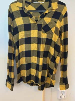 Gul och svart rutig skjorta H&M - Snygg långärmad skjorta från H&M i gult och svart rutigt mönster. Skjortan har klassisk krage, bröstficka och knäppning framtill. Perfekt för en avslappnad och trendig stil. Storlek 164 barn.