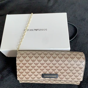 Emporio Armani - Emporio Armani väska i beige/brun färg med Armanimärken. Medföljer en guldkedja med svart läder som man spänner fast inuti väskan men den kan tas bort om man hellre vill ha den som en plånbok. Finns fack för kort inuti samt ett fack med dragkedja. 