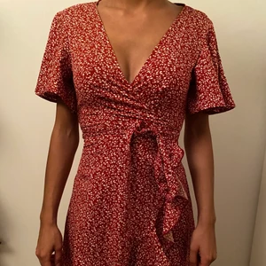 Röd blommig klänning - En röd omlottsklänning med vita blommor som ett mönster. Den har två knytband som man knyter tillsammans runt midjan. Denna är i storlek XS. Köpt ifrån SHEIN och säljes för 50kr👗