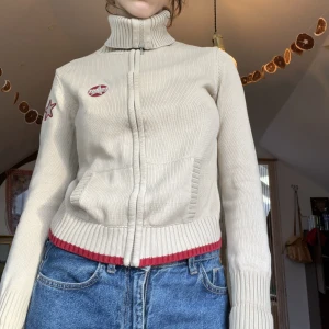 Esprit zipup - Beige zip-up från Esprit med röda stjärn detaljer på armen och bröstet! Lång krage som kan sylas påde öppen och stängd.