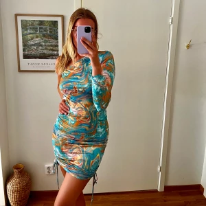 Mönstrad klänning i blått - Oanvänd klännig i en fin blå mönstrad färg med inslag av andra färger såsom orange. Går att göra kortare samt längre med snörena på sidan. Stretchig så passar större och mindre strl också.