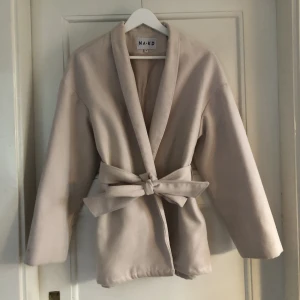 Nakd kappa - Beige/ljusrosa kort kappa med bälte från nakd, kanten nertill var avklippt och uppsydd när jag köpte den (bild 3) men det är inget som märks när man använder den. Bra skick förutom en liten fläck på ena armen, passar till allt! Lite oversize så passar s/m