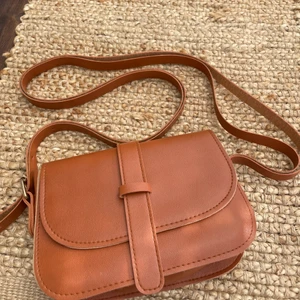 Brun axelremsväska - Säljer en brun/ läderliknande axelremsväska som aldrig har kommit till användning. Den är i nyskick, säljer för 50kr + frakt