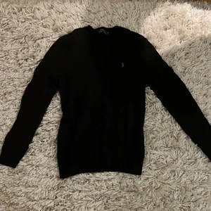 Ralph Lauren Stickad Tröja - Stretchigt material, passar månag kroppstyper. Stickad svart tröja med en lila Ralph Lauren logotyp. Inköpspris 1299kr.