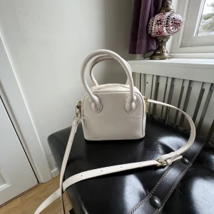 Väskor - En beige axelväska med gulddetaljer. Den är i gott skick och är dessutom rymlig. Axelbandet är både justerbart och möjlig att ta av