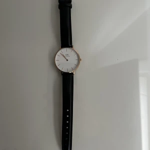 Daniel Wellington 34mm - Jättefin och knappt använd DW 34mm med svart läderband och boett i rosé. Tillkommer även ett helt oanvänt läderband i brun färg som är i oöppnad förpackning. Säljer pga present som inte kommit till användning. 