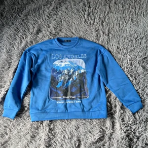 Blå sweatshirt med Los Angeles-tryck från SHEIN - Blå sweatshirt från SHEIN med stort tryck av berg, skog och texten 'Los Angeles', 'World Peace' och 'Happy Every Day' på framsidan. Rund halsringning och långa ärmar. Perfekt för dig som gillar streetstyle och coola prints. Storlek M