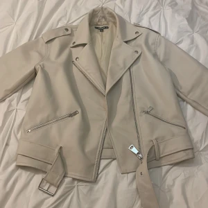 Beige skinnjacka - Beige skinnjacka från Nelly i mycket fint skick utvändigt, finns dock sminkfläckar på insidan av kragen och svarta fläckar längst ned på insidan (färgat av sig från jeans) men det är ingenting som syns när man har den på 🙌 Bältet kan man ta av om man vill