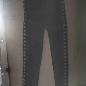  - Säljer ett par Kappa-byxor. Dom är köpta i Köpenhamn förra året och är knappt använda. Byxorna har en revär med kappa-märket och har även kappa-märket på höger lår. Byxorna har knytning i midjan. Ganska lågmidjade 