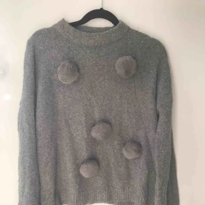  - Säljer en tröja köpt på Benetton. Tröjan är stickad och har ett par fluffiga bollar på framsidan av tröjan. Det är en mörkgrå tröja.