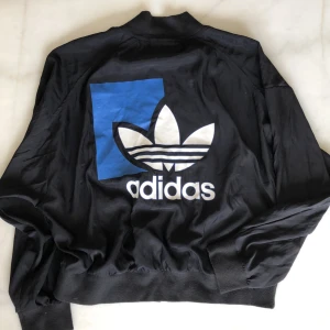 Adidas jacka  - Adidas jacka i tunt material med stort tryck på ryggen. Oversize modell så passar flera storlekar. Använt men fint skick. Frakten är spårbar och inräknad i priset