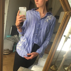  - Ljusblå Linneskjorta med smala ränder och blommorna på höger axel/bröst samt vänster höft