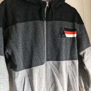 Le Coq Sportif-hoodie - Grårandig huvtröja med ett litet tryck frampå bröstet och ett stort tryck baktill. Storlek S. 