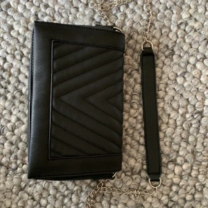 Läderväska med kedja - En svart läderväska från PIECES som är köpt på Vero Moda med mönster på framsidan, kedjor-band och ett fack med blixtlås inuti. Använd få gånger & bra skick! Köparen betalar frakten (;