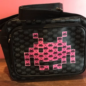 Space invaders äxelväska - Helt ny väska med retro spelet Space Invaders motiv. Svart väska med rosa motiv i storlek 30*24*10cm. Märket på väskan är SkyLine. Axelremsväska som kan hämtas upp i Enskededalen eller skickas mot fraktkostnad.