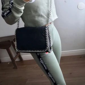  - Svart axelremsväska i skinnimitation med kantad silverlänk. Ett fack med dragkedja på baksidan och ett fack med dragkedja på insidan. Axellänk 105 cm. Längd 28 cm, höjd 18 cm, bredd 5 cm.  Köpt på Scorett för 499kr. Sparsamt använd, bra skick!