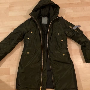 Grön vinterjacka från Alpha industries  - Säljer min snygga mörkgröna vinterjacka från Alpha industries. Den skimrar lite i metallic grönt och har guldfärgade detaljer så som knappar och dragkedjor. Dragkedjan kan dras ned uppifrån men även underifrån (dubbla dragkedjor).                    Den är köpt förra året på Åhléns och är som ny, INGA defekter, storlek S. Nypris 1999 - mitt pris 450 inklusive frakt. Kan även mötas omkring Hökarängen och och isf är priset 400kr 🥰