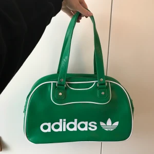 Grön väska från Adidas, helt oanvänd! - Så snygg grön väska från Adidas (äkta), den är helt oanvänd så den är i nyskick! Mått: längd 37 cm, bredd 13 cm, höjd 22 cm, handtagens längd ca 24 cm. Inuti väskan finns tre fack samt ett fack med dragkedja. Vid frågor är det bara att skicka iväg en kommentar eller meddelande💚