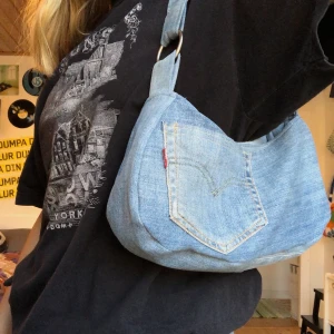 Handsydd handväska jeans  - En jeans väska i Inga modell med Levis ficka på ena sidan, silver ringar och ljusblått foder. Den har som vanligt dragkedjsficka i matchande färg samt tryckknapp. Frakt:44kr 