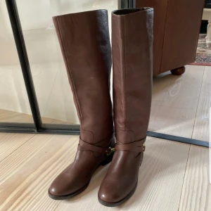  - Fantastisk stövlar från Ralph Lauren i ridmodell. Mörkbrunt läder med guldsporre och detaljer i guld. Smala i skaftet. Aldrig använda. Omkrets 36 cm, höjd (totalt) 42 cm. Storlek 37.5