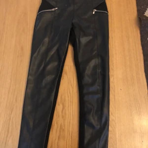  Svarta skinnbyxor (fake) stl 128 - Supersnygga svarta skinnbyxor (fake), ”skinn” på framsidan, stretchigt tyg på baksidan (se bild). Byxorna är från Mayoral i stl 128. De är i använt skick, därför så lågt pris.