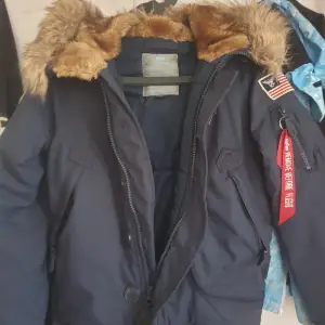 Snygg jacka med massa fina detaljer från Alpha Industries. - Luva med avtagbar fuskpäls - Stängs med dragkedja och knappar - Femficksmodell arav en invändig - Ribbstickad mudd ärmslut och nederkant- - Fodrad