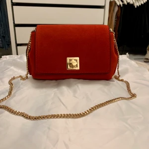 Röd väska i mocka från Mango  - NY. Supersnygg röd väska i mockaimitation från Mango med guldkedja. Perfekta väskan i vinter! Tyvärr har den bara blivit ståendes i min garderob då jag har så många väskor och söker nu nytt hem. Aldrig använd! Rymlig med fack inuti. 24x14 cm. 