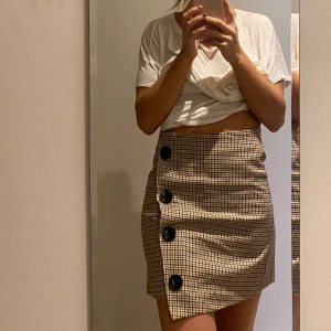 Kjol  - Kjol i små rutor med fin knapp-detalj längs sidan! Dragkedja. Köpare står för frakt.