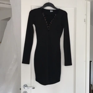  - Klänning med lång ärm, svart blank ribbad  Tight fodralklänning med roséfärgade öljetter och snörning fram Mycket fint skick - använd en gång! 