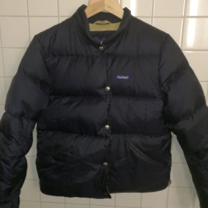  - Varm och snygg dunjacka i bomber-modell från Penfield. Jackan är marinblå på utsidan och beige på insidan. Gott begagnat skick. En söm i ena ärmen har släppt (se bild), går enkelt att laga hos skräddare. Strl S. Fraktkostnad tillkommer. 
