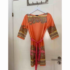  - Orange mönstrad klänning med knytbart band i midjan, kan även knytas bakom ryggen så klänningen ser ut att sitta tightare. Storlek 36. Frakt tillkommer.