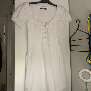Vit klänning från Bik Bok - Säljer en superfin vit klänning från Bik Bok i storlek XS. Klänningen har korta puffärmar och en snygg snörning framtill. Den är perfekt för sommarens alla fester och har en normal passform. Klänningen är i nyskick och har en dragkedja på sidan för enkel på- och avtagning. Passar perfekt till både vardag och fest! Man kan även skjustera storleken fram med banden 🎀