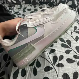 Säljer ett par Nike Air Force 1 Shadow sneakers i flera färger. Skorna har en vit bas med grå, rosa och gröna detaljer. De är tillverkade i skinn och syntetmaterial och har en normal passform. Perfekta för både vardag och träning!