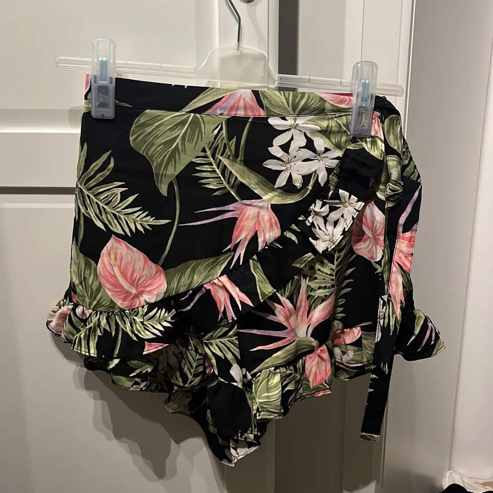 En färgglad kortkjol från BikBok med ett blommigt mönster i grönt, rosa och vitt på en svart bakgrund. Kjolen har volangdetaljer och ett knytband i midjan.. Hameet.
