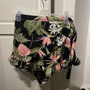 Blommig kortkjol från BikBok - En färgglad kortkjol från BikBok med ett blommigt mönster i grönt, rosa och vitt på en svart bakgrund. Kjolen har volangdetaljer och ett knytband i midjan.