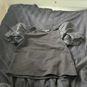 Svart topp med puffärmar från KappAhl - Säljer en svart topp från KappAhl i storlek 146/152. Toppen har puffiga ärmar i ett glansigt material som ger en snygg kontrast till den matta kroppen. Perfekt för både vardag och fest!