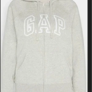 Grå hoodie från GAP - Säljer en supermysig grå hoodie från GAP. Den har en dragkedja framtill och en stor GAP-logga på bröstet. Perfekt för höst och vinter när man vill hålla sig varm och bekväm. Den är i bra skick och passar perfekt till vardags eller chillkvällar. Köp den nu eller ge ett bud! 💛