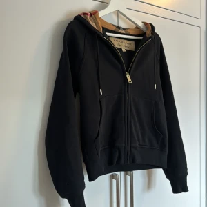 Svart hoodie från Burberry - Säljer en snygg svart hoodie från Burberry. Den har en dragkedja framtill och en klassisk Burberry-rutig insida på huvan. Perfekt för höst och vår. Hoodien är i nyskick och har långa ärmar. Passar perfekt för en casual look eller när du vill vara bekväm men ändå stilren. nypris runt 4000