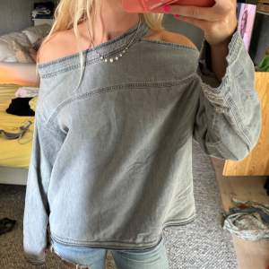 Säljer en snygg grå off-shoulder tröja i jeansmaterial. Tröjan har en cool asymmetrisk design med en axel bar och en lång ärm. Den är i jättebra skick och aldrig använd, endast testad! 