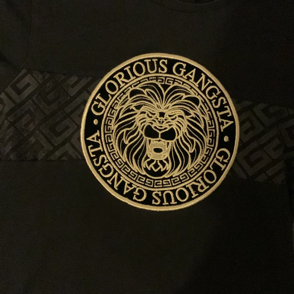 Säljer en svart t-shirt från Glorious Gangsta i storlek S. T-shirten har ett stort, guldigt lejontryck på framsidan med texten 'Glorious Gangsta' runt om. Den har också ett mönster i svart och guld på ärmarna och över bröstet. Perfekt för en cool och avslappnad stil!. T-shirts.