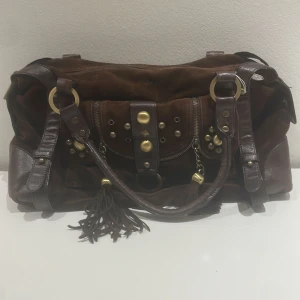 Brun axelväska med fransar och nitar - Säljer en snygg brun axelväska i skinn och mocka. Väskan har coola detaljer som fransar och nitar i guld. Den har en dragkedja och flera fickor för förvaring. Perfekt för både vardag och fest!