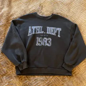 Säljer en mörkgrå sweatshirt med trycket 'ATHL. DEPT 1983' på framsidan. Tröjan är i normal passform och har långa ärmar. Perfekt för en casual look eller för att mysa hemma. Den är i bra skick och väldigt bekväm att ha på sig. köptes för runt 300kr men säljs för 30kr.